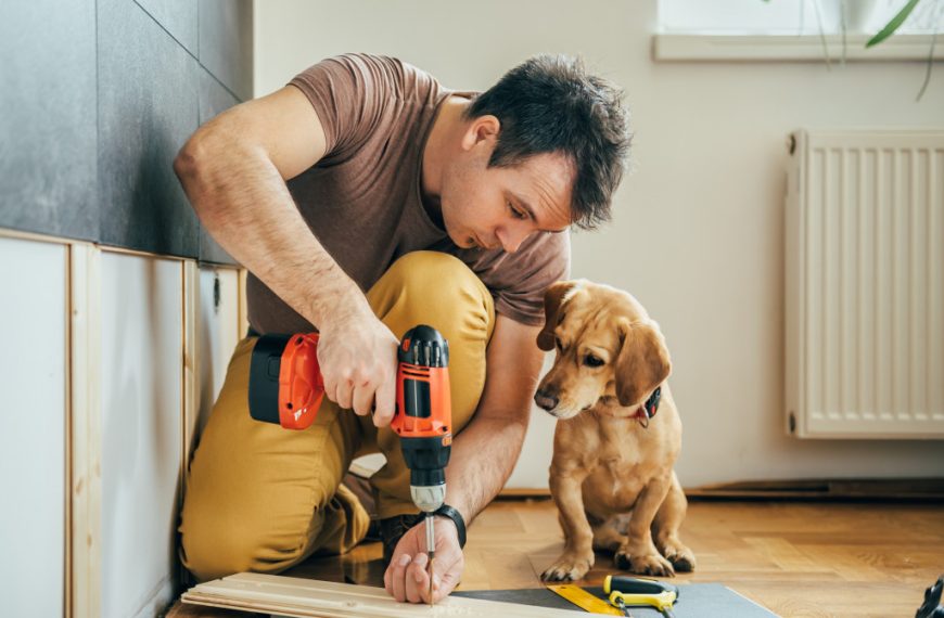 A man using a hand drill on wood while his dog watches
